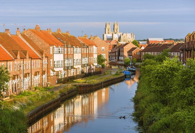 houses in a row adjacent to a canal, 2 narrow boats are in the background of the image, the houses reflect into the water.