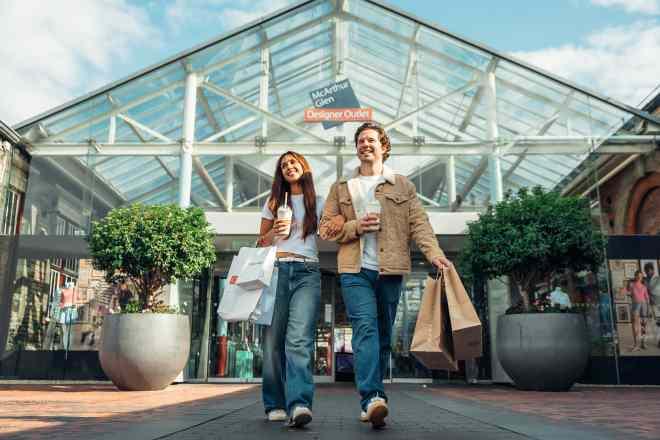 "Two people walking out of the Swindon Designer Outlet entrance under a glass canopy, carrying shopping bags and drinks, with large planters on either side of the doorway"
