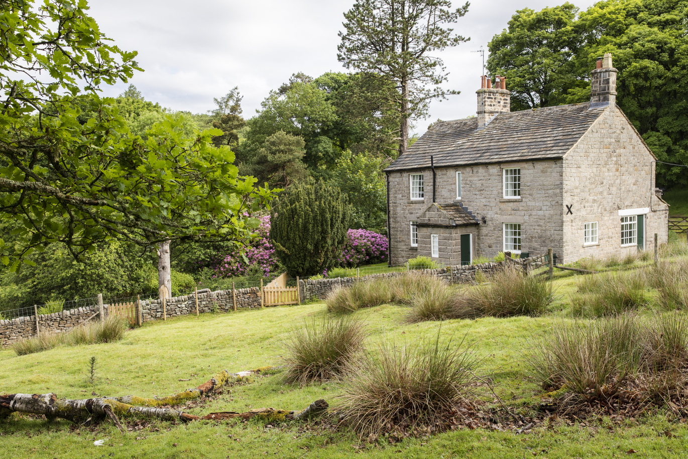 The exterior of Yarncliff Lodge, a stone building surrounded by lots of trees, plants and flowers in a lush green lawn with a brick wall down the centre, dividing the lawn and another field/lawn.