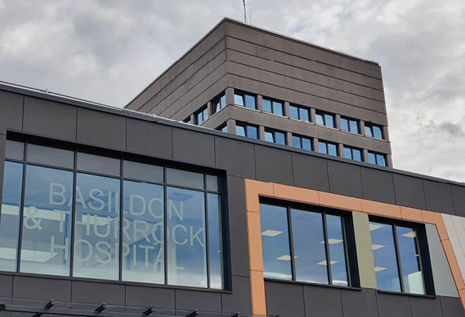 "Close up of upper levels of Basildon University Hospital. The building has black cladding and has a sign that reads ' Basildon and Thurrock Hospital' in the window. There are grey clouds in the background"