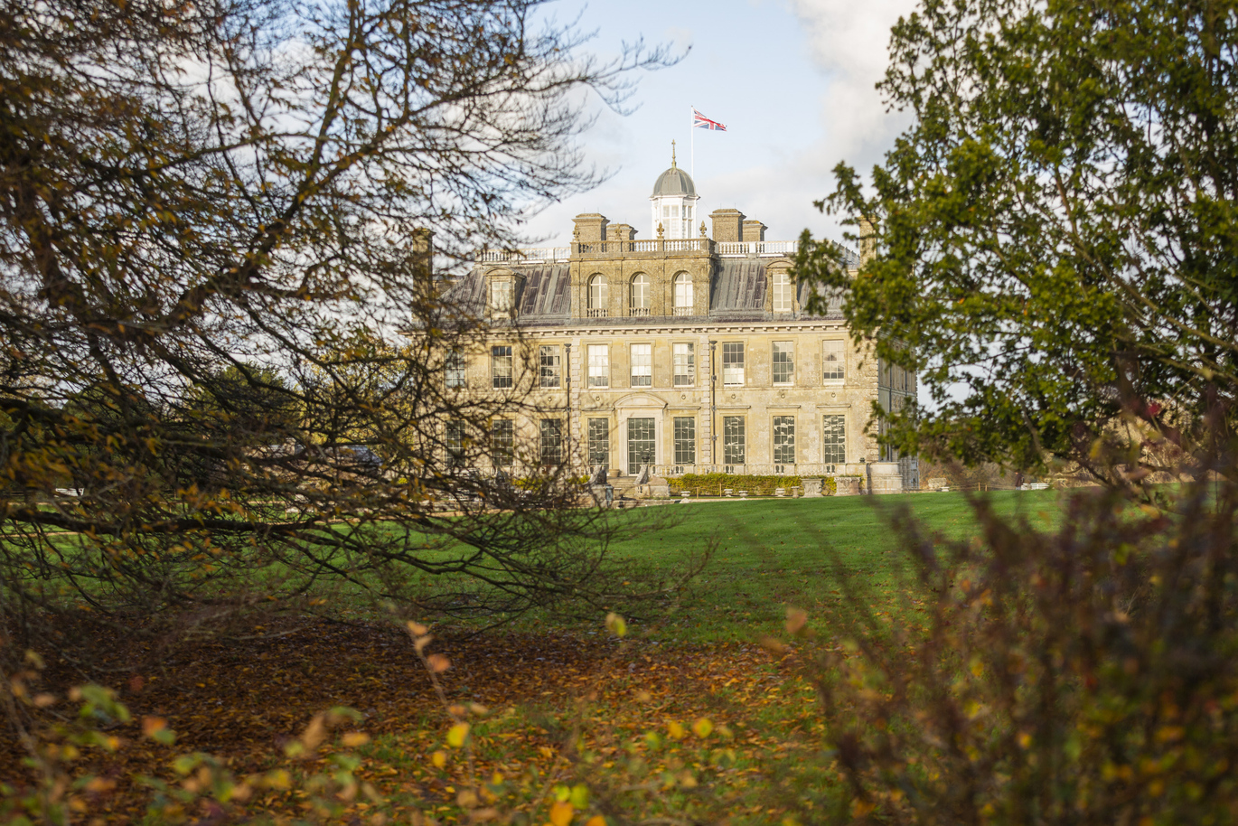 View through the trees of the large house through the park at Kingston Lacy with a lush green lawn out the front scattered with autumnal orange leaves.
