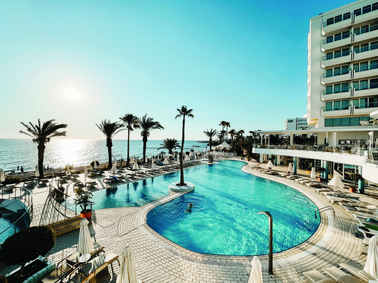 Aerial view of the pool area with one single person having a swim with sun loungers and umbrellas surrounding. With part of the hotel building to the right and the sea to the left with the sun rising above.