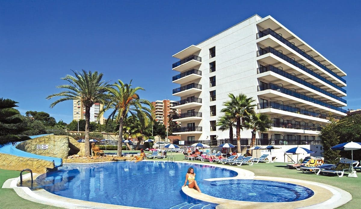 View of the pool area with a water slide, surrounded by deck chairs and umbrellas with palm trees around. With the tall hotel building behind on a clear sunny day.