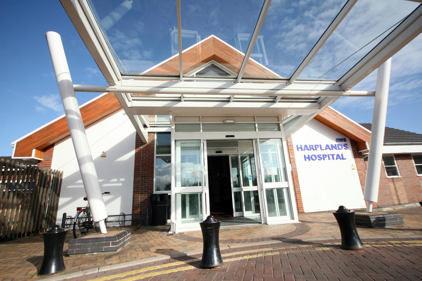 "Main entrance to Harplands Hospital. In front of the entrance is a glass roof canopy. There are 3 bollards on the road just outside the entrance".