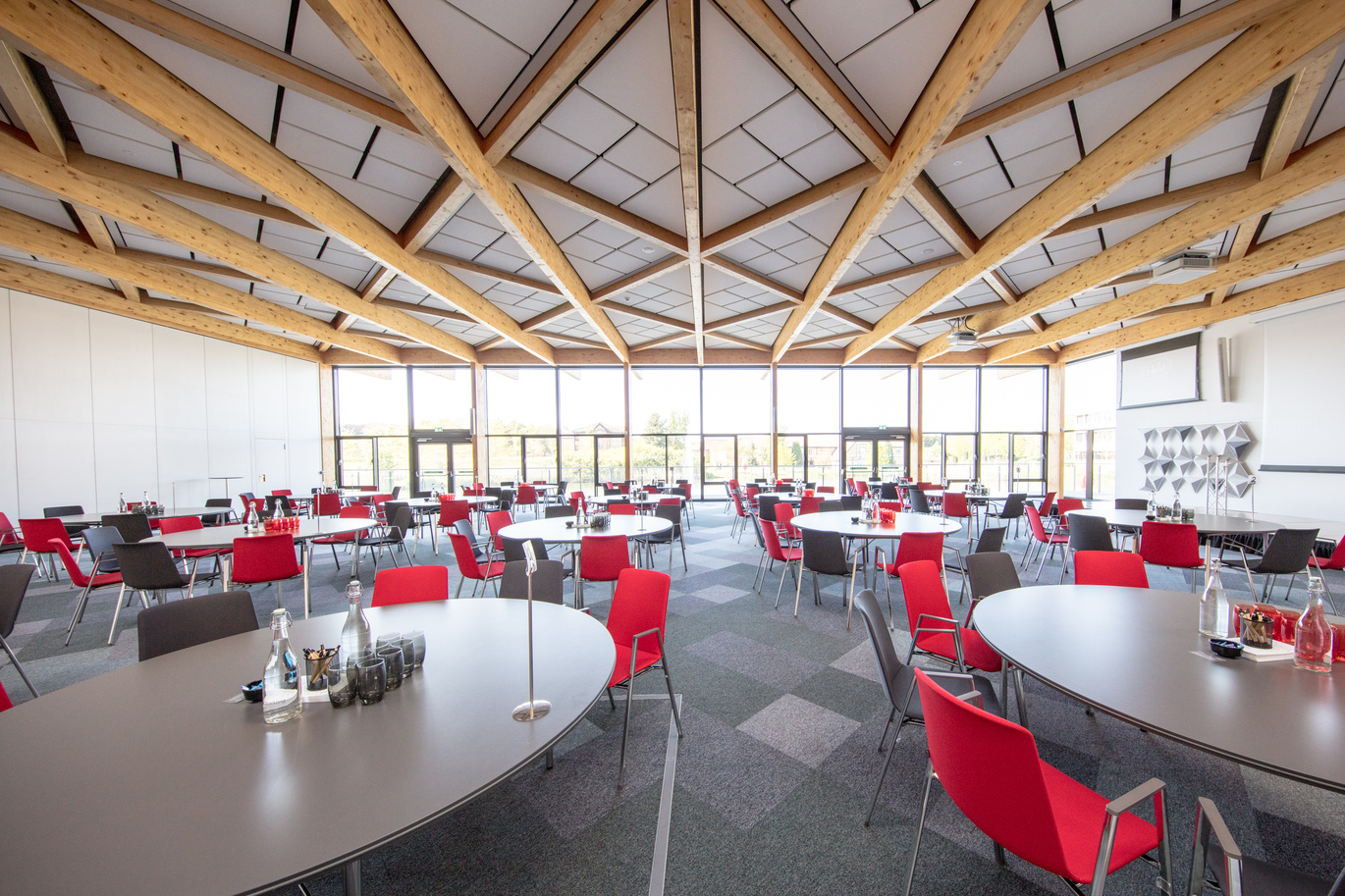 "Large Conference room with large round grey tables, alternating red and grey backed chairs, High Ceiling with wooden rafters and full height floor to ceiling windows at the end of the room"