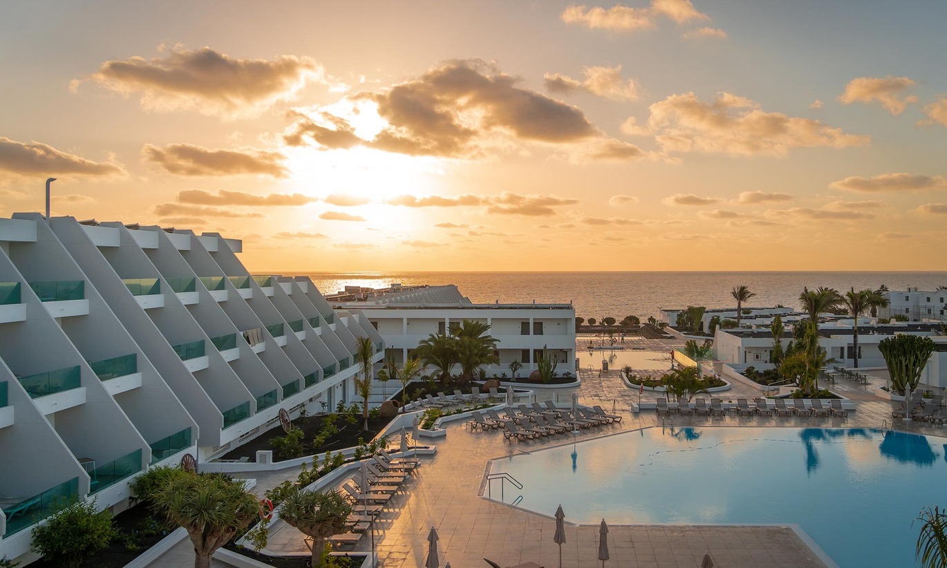 View of the hotel and pool area with neatly places sun loungers around the pool with the sea behind with the sun setting above.
