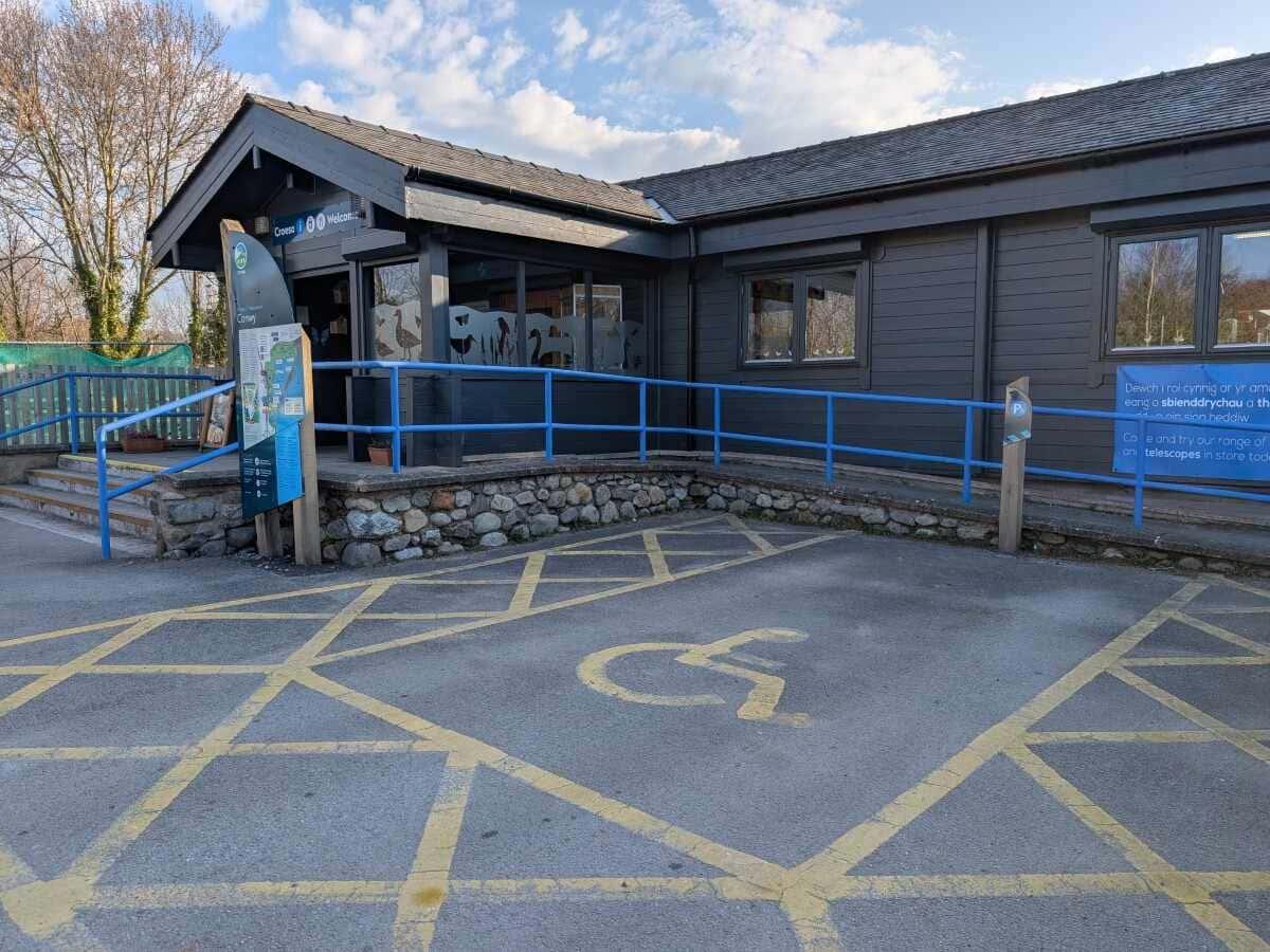 Accessible entrance to a visitor centre featuring a ramp and informative signage, surrounded by trees and blue skies
