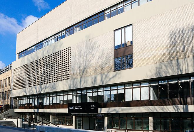 “Exterior view of Camberwell College of Arts Peckham Road campus, featuring a modern multi-story building with light-colored brickwork, large horizontal windows, and a patterned brick section on the upper facade. A black sign with the UAL logo is visible above the entrance, and tree shadows fall across the building.”