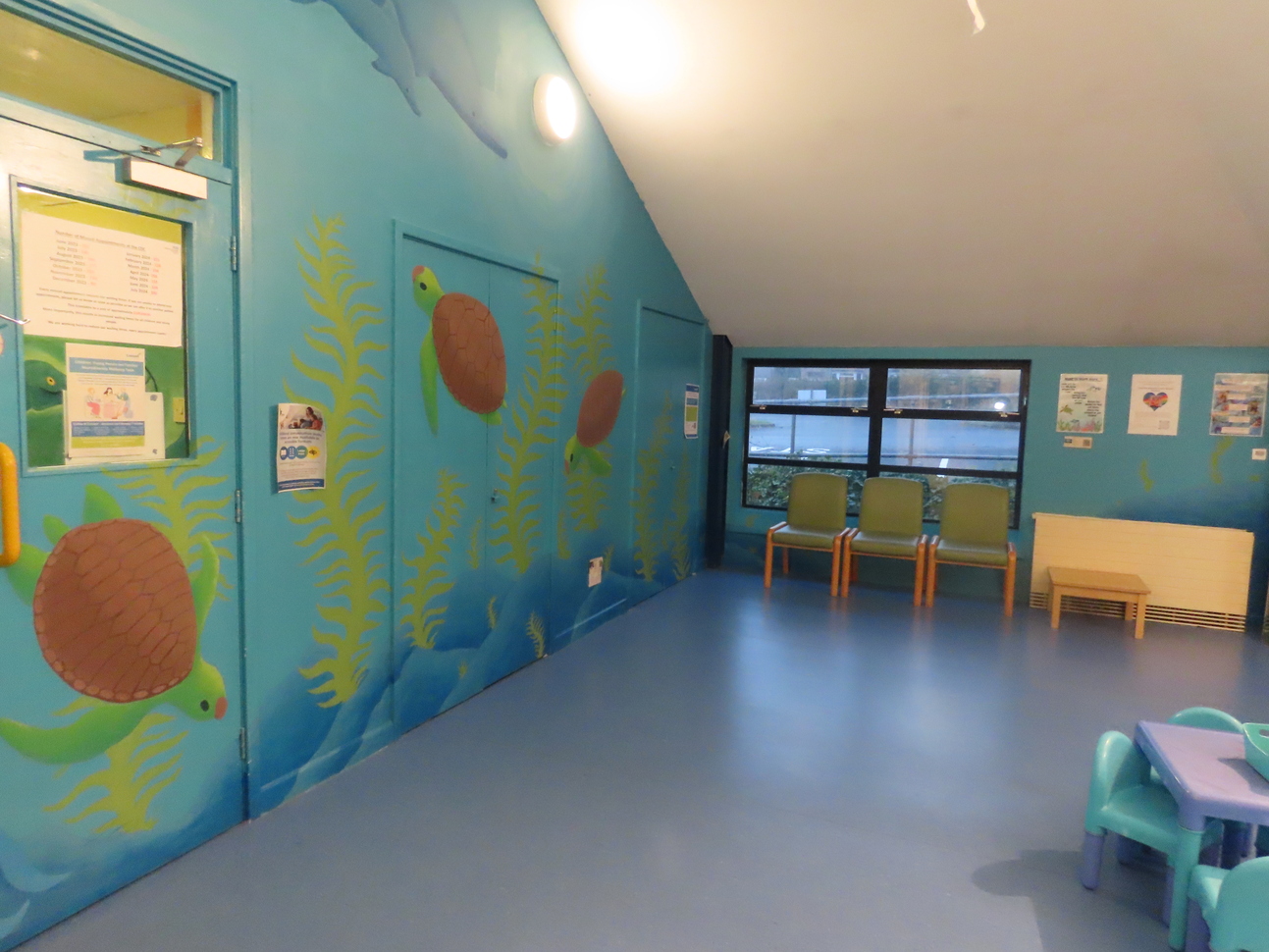 A colorful waiting area with an ocean-themed design. The walls are painted blue with illustrations of sea turtles and underwater plants. The room includes a window overlooking the outside, four green chairs lined up against the wall, and a small wooden table. In the foreground, a child-sized plastic table and chairs are visible