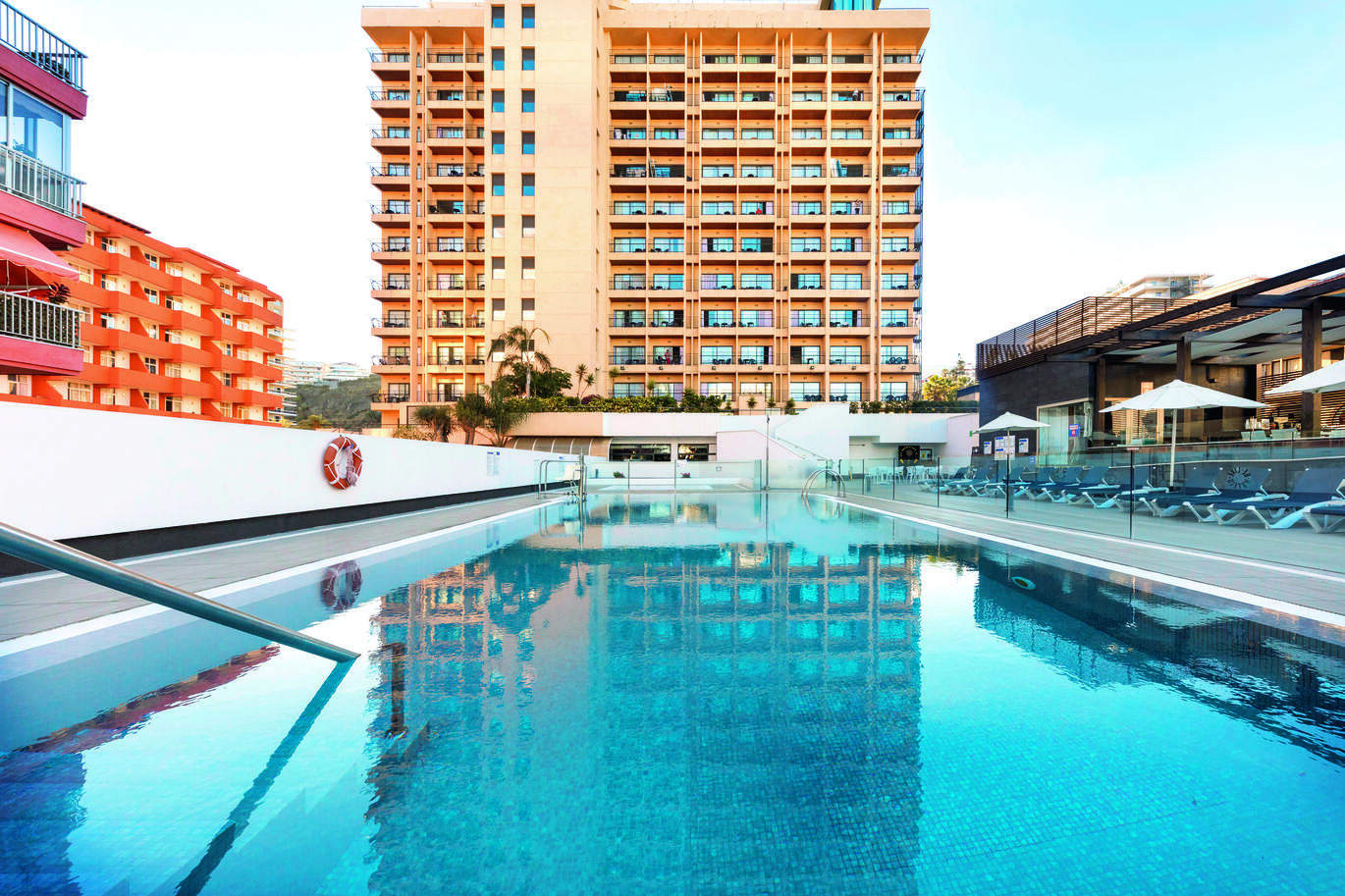 View from the pool of the tall hotel building behind. With glass panels at the edge of the pool and deck chairs with umbrellas behind.