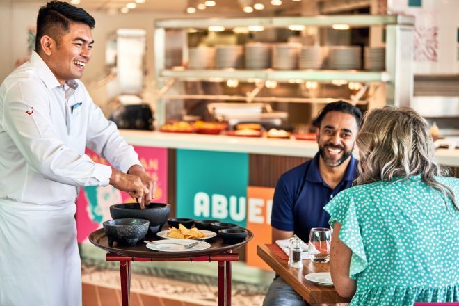 "A member of staff serving food to a couple sitting opposite each other at a table in a restaurant. The restaurant counter can be seen in the background with plates stacked up on a shelf and prepared food beneath them."