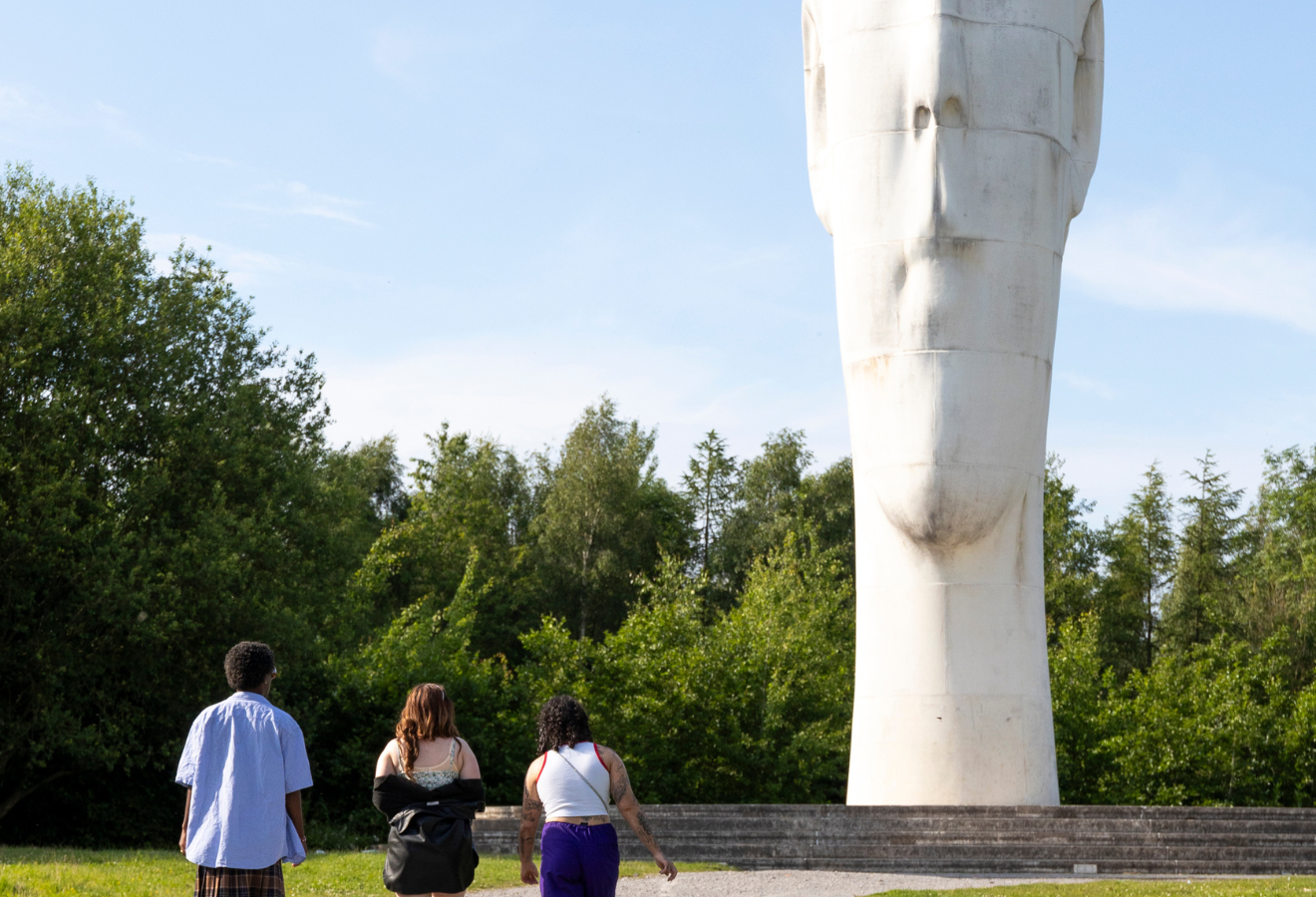 3 people stand in front of a white statue, with thier backs to the camera admiring the large face, trees are in the background 