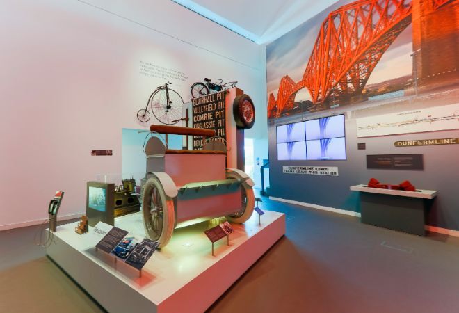 "Exhibit displaying transport items. Penny farthing on wall in background. Picture of the Forth Road Bridge in background to right with 4 screens underneath."