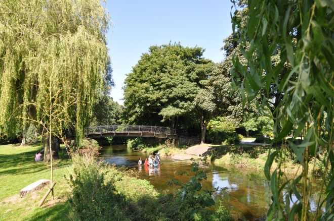 "photo of King's park Retford. A river goes through the park, with green covered banks on either side. a family plays in the river, and a bridge crosses in the background"