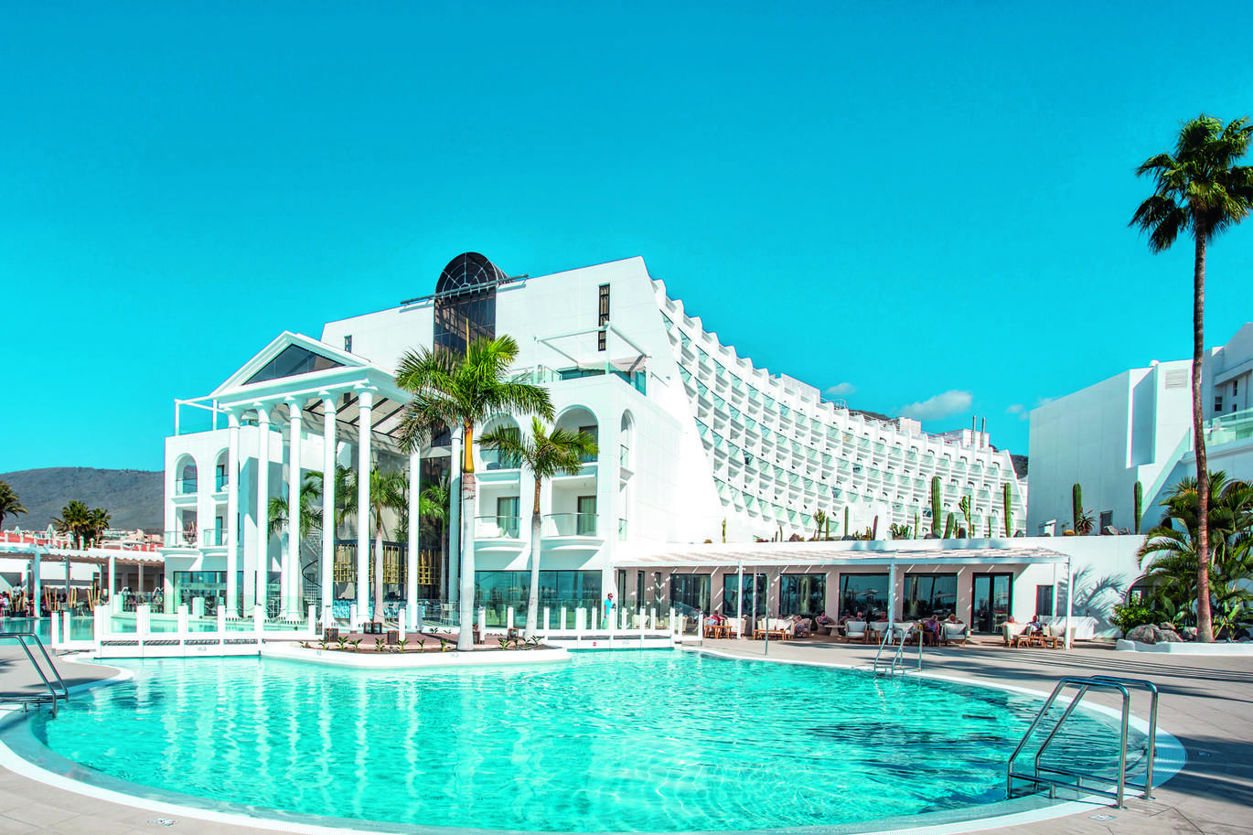 A view of the large, white, elegant hotel building with the lush blue pool out the front with some undercover seating in between the hotel and pool. There is a small white bridge through the middle of the pool and a few palm trees around. It is a lovely clear blue day.