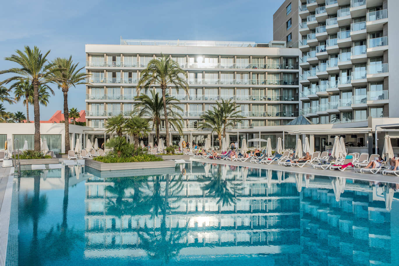 View of the pool with people on sun loungers with umbrellas surrounding the pool with palm trees dotted around. Behind is the large hotel building with clusters of hotel room balconies on a sunny day.