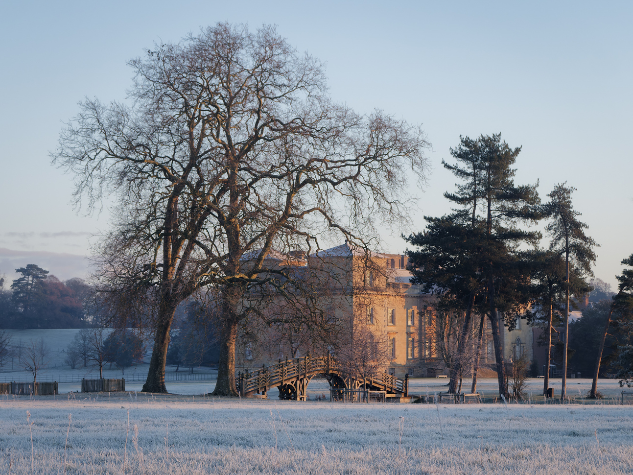 A large medieval type building in the middle of a frosty field surrounded by more fields and trees with a Chinese type bridge in front also surrounded by trees.