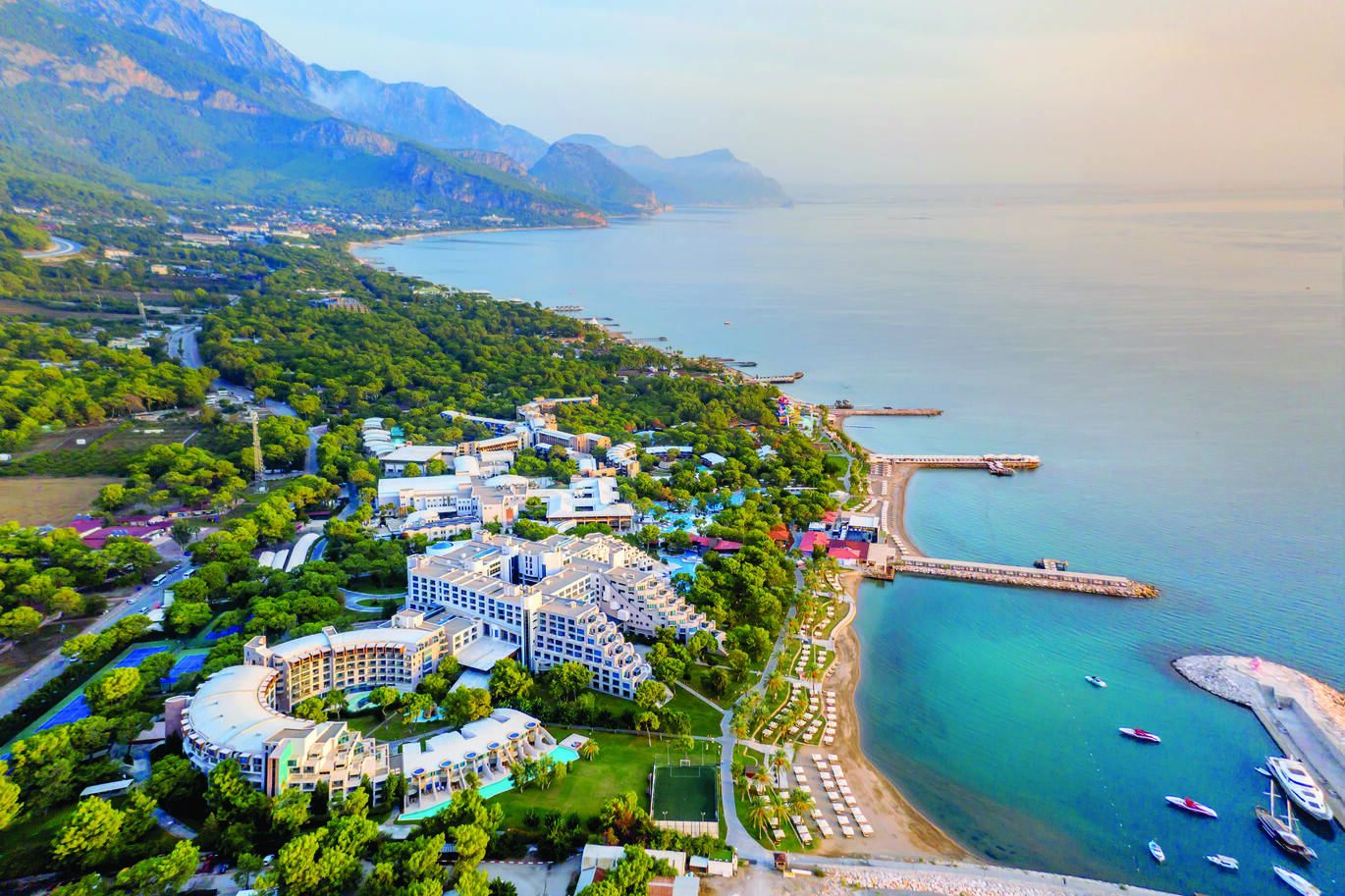 Aerial view of the hotel complex right on the beach, surrounded by many trees and big mountains in the background.
