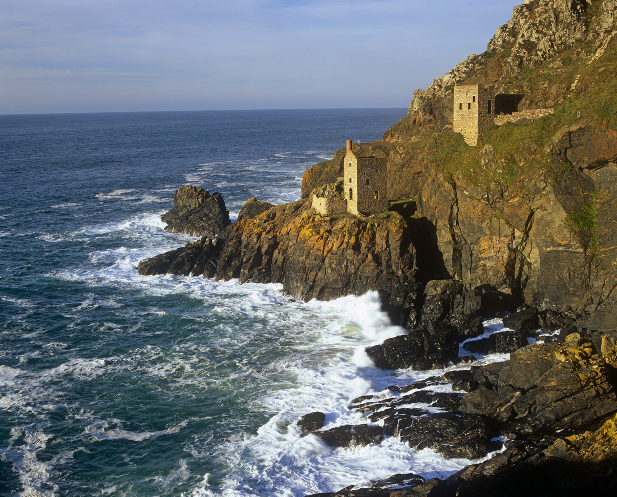 The abandoned beam engine houses of the Crowns section of Botallack Mine on the edge of a cliff above the sea with waves crashing below.