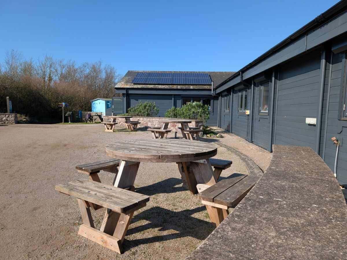 Outdoor seating area with wooden picnic tables near a building with solar panels under a clear blue sky