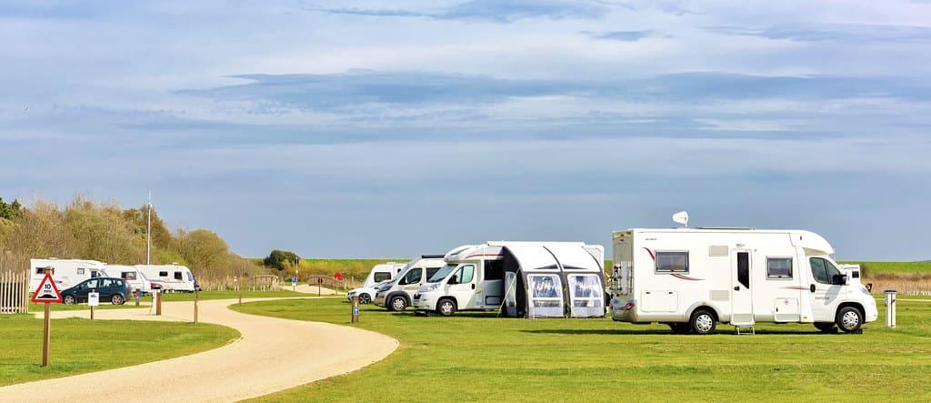 A serene campsite with several parked RVs and motorhomes on a green grass field. A winding path leads through the area under a clear, blue sky.