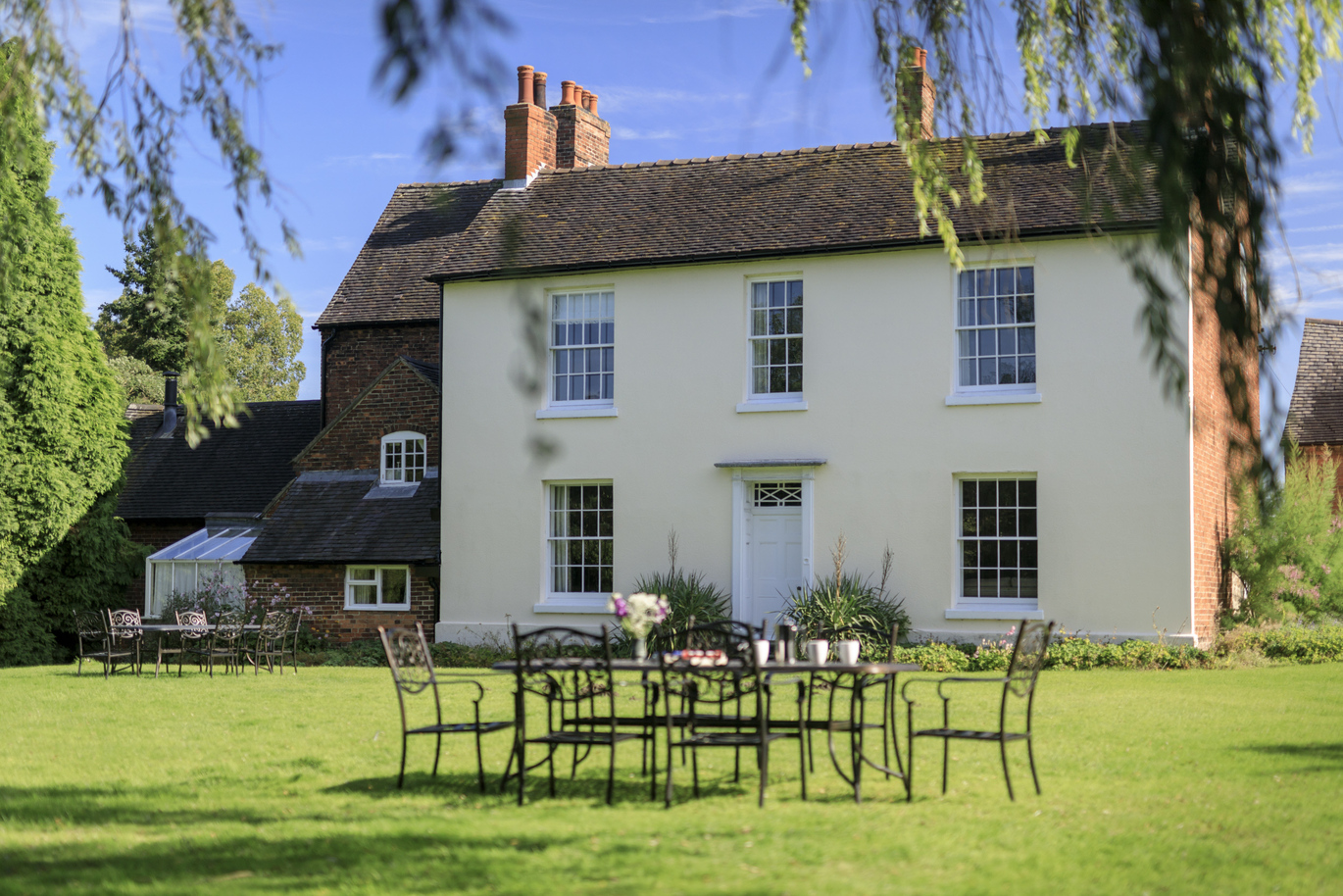 The exterior of Southwood House Farm, the front of the building is magnolia while the side and other parts of the building are brick. There are 2 large chimneys on top of the building and a small conservatory on the left side of the building. At the front is a very green lawn with some plants at the foot of the building and 2 large tables with chairs on the lawn.