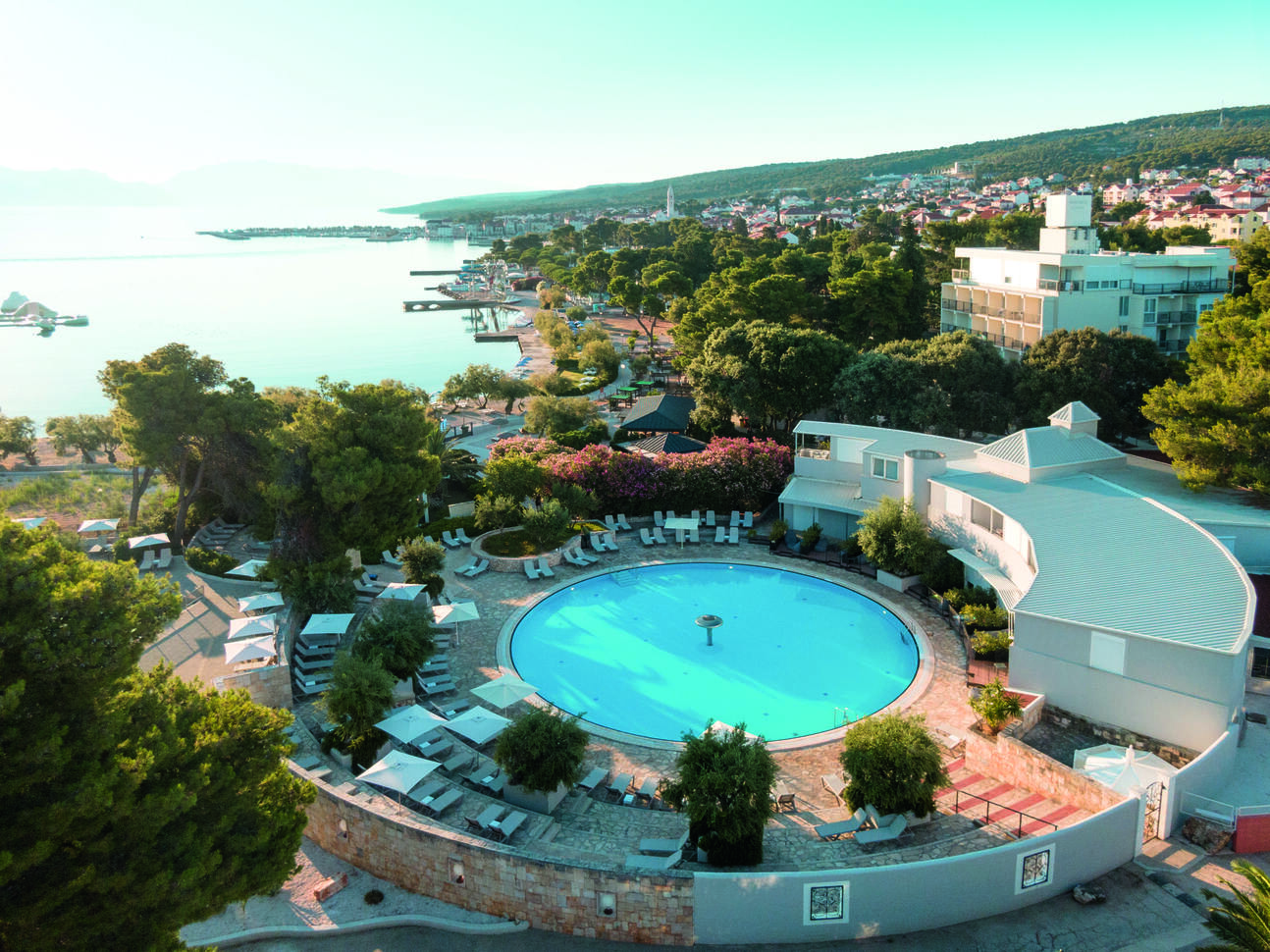 Aerial view of the round pool area surrounded by sun loungers and part of the hotel building. With the sea and other areas in the background.