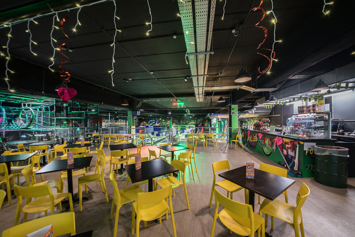 "Restaurant area with yellow tables and black chairs. There is a counter to the right of the area. There are fairy lights from the ceiling"