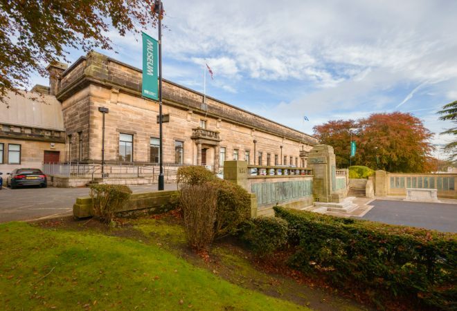 "Kircaldy Gardens. Sandstone building. Garden area with neat hedges and courtyard in foreground. Autumnal trees in background set against blue cloudy skies."