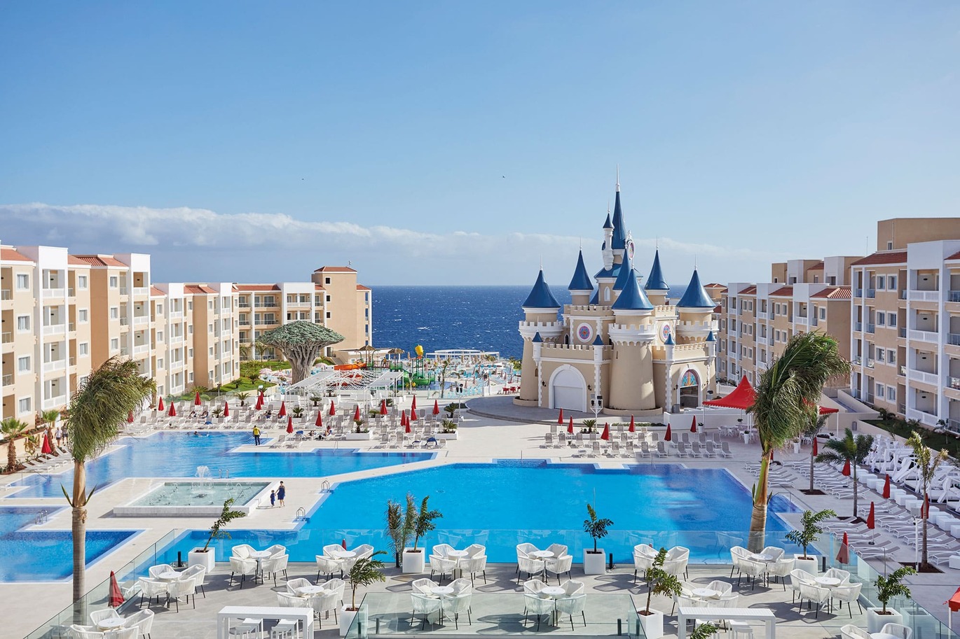 A view of the 2 large and 1 smaller swimming pools with a water fountain feature in the middle and number of palm trees dotted around. The pools are surrounded by white sun loungers with red collapsed umbrellas. The hotel buildings are surrounding either side with a large castle building in the middle behind the pools. At the front of the image is a seating area with white tables and chairs bordered with glass. You can see the sea behind with the blue, slightly cloudy sky above.