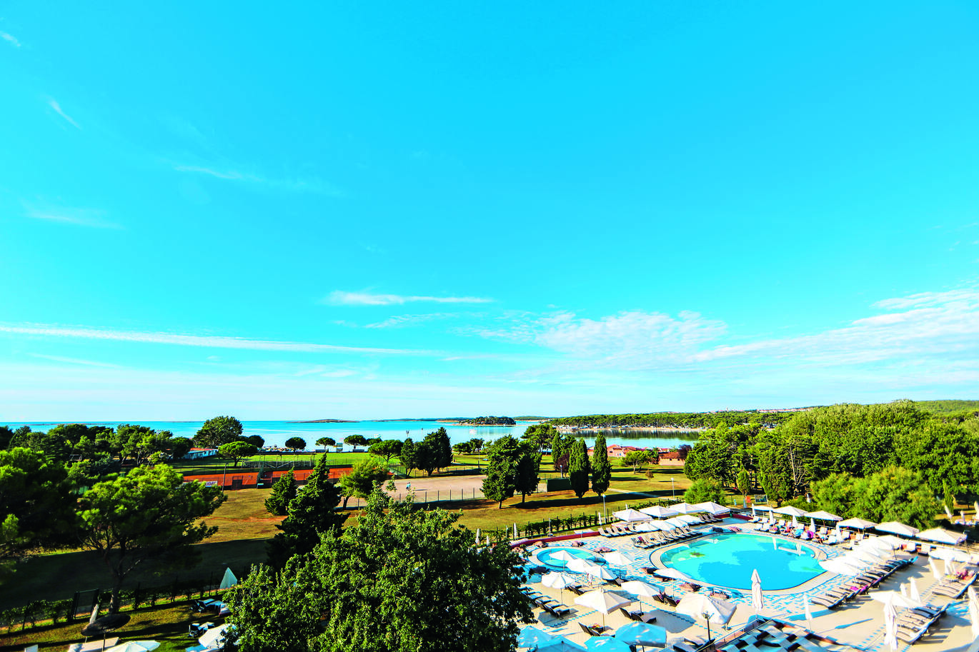 View of the hotel pool surrounded by sun beds and umbrellas with many trees surrounding the area. There are two courts beyond and the sea beyond that.