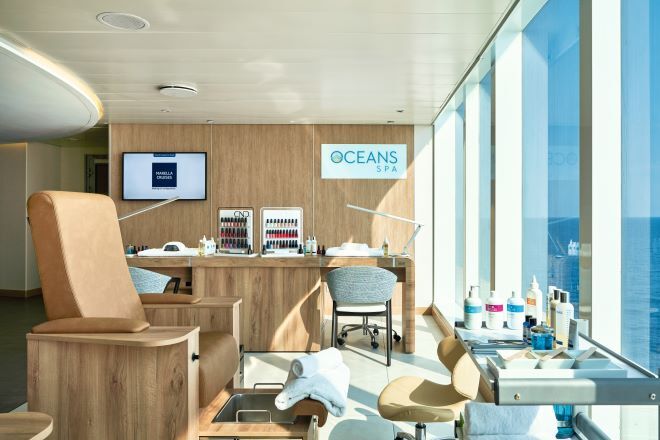 "Oceans Spa onboard Marella Voyager. In the foreground there is salon chair with a foot spa underneath next to a table with beauty products on. In the background are two tables for nail painting. There is an array of different colour nail polishes on the back wall as well as a TV and sign that reads 'Oceans Spa'."