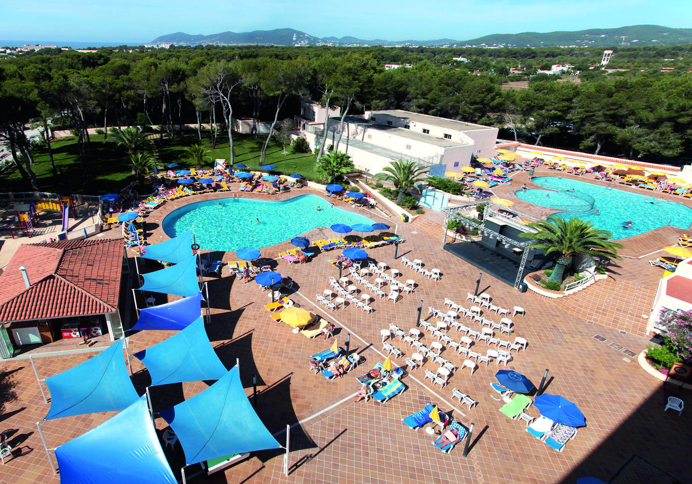 Aerial view of the pool area with two pools surrounded by sun loungers and many trees behind.