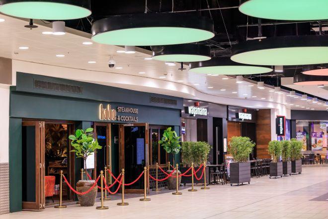 "Restaurant frontage at Xscape with outdoor seating, potted plants and rope barriers along a wide indoor walkway."