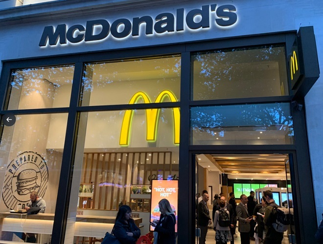 "Photo of a McDonald's entrance taken from outside showing people inside standing. There is a large 'M' symbol located at the top of the wall inside. There is seating to the left. The entrance door is to the right and is open."