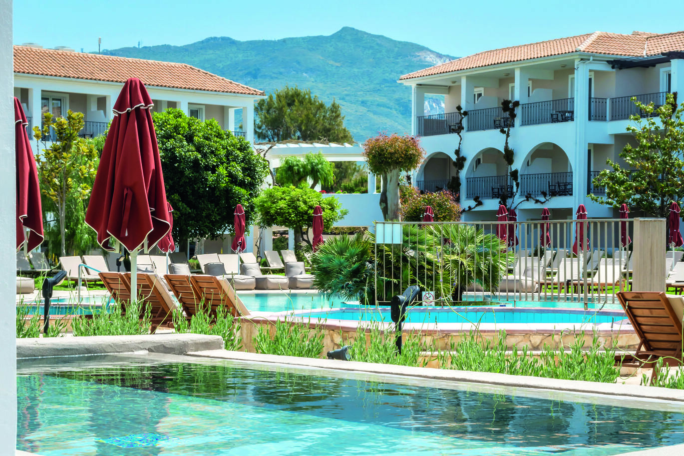 View of the pool area surrounded by sun loungers and bean bags with trees all around and clusters of hotel rooms behind, on a clear, sunny day with a mountain in the background.