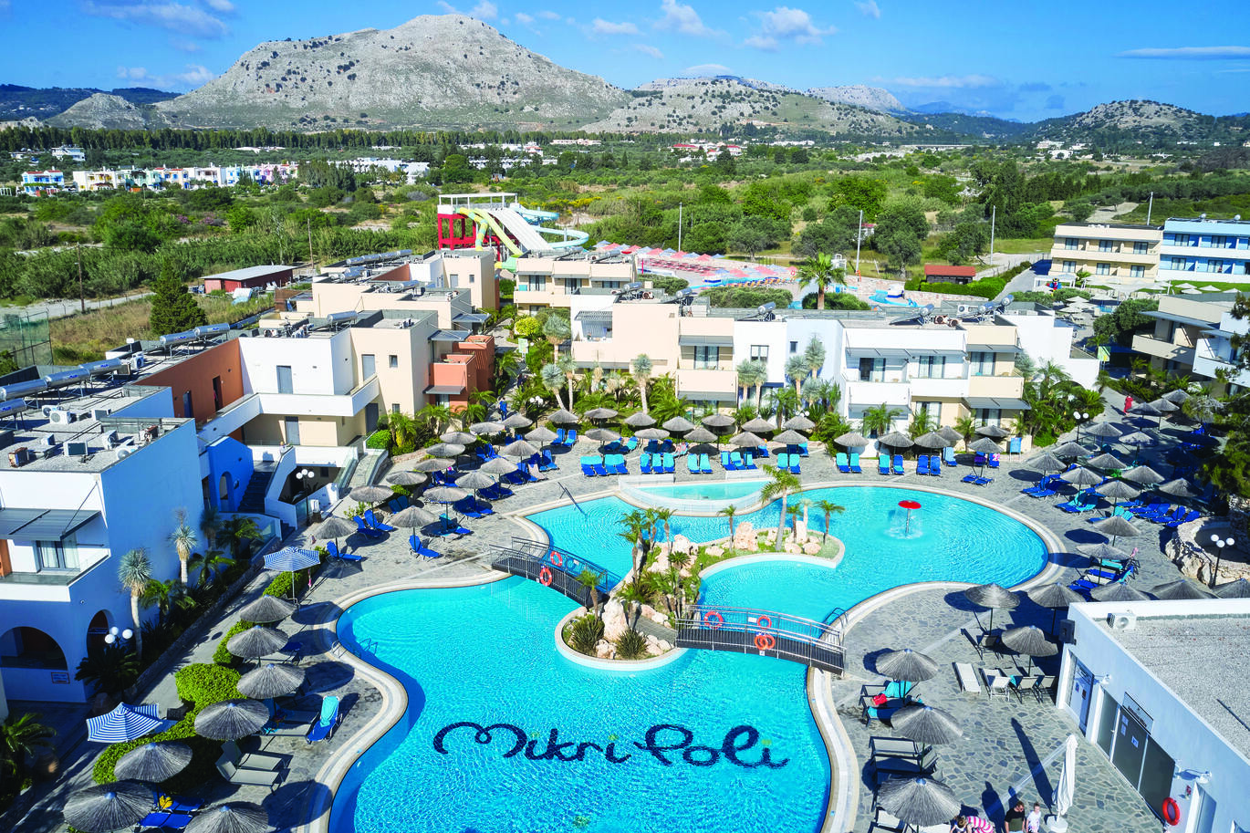 Aerial view of the hotel complex with with trees. buildings and large hills in the background. With the pool in the middle with a small island with trees on in the middle of the pool with a path and two bridges to get from one side of the pool to the other. Sun loungers with umbrellas surround the pool with the hotel buildings surrounding and tall water slides behind the buildings.
