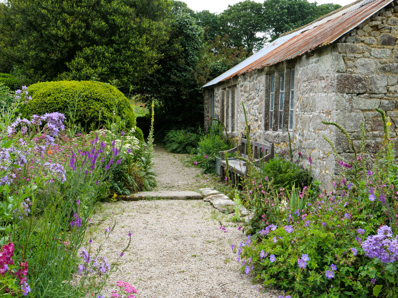 An old stone outbuilding with a wooden bench up against the exterior with a stone path surrounded by many colourful plants and lots of greenery.