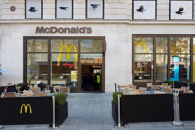 "Photo of outside of a McDonald's. There is tabled seating either side of a pathway to the main entrance door. There are black barriers surrounding the seating area to close off from the street. There is a McDonald's sign above the entrance doors. Some people are queuing inside the entrance and some people are seated outside."