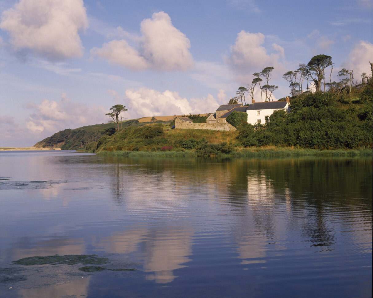 A view over the lake of the farm house on a hill of grass and trees.