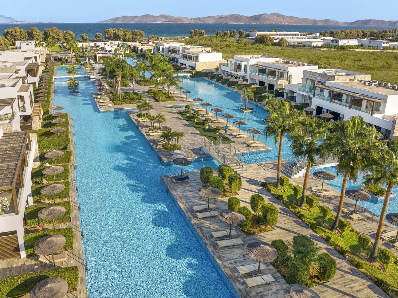 Aerial view of the hotel with pools surrounding and the sea behind with mountains beyond.