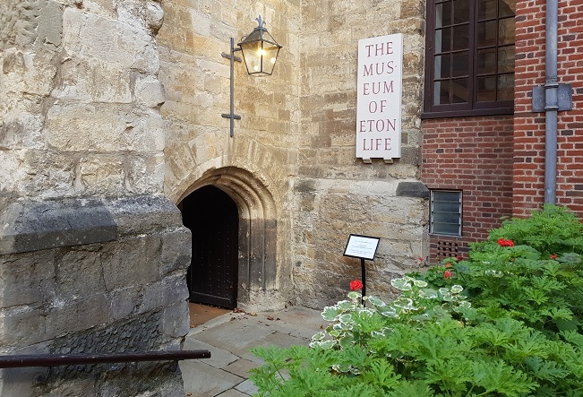"Photo of Museum of Eton Life. There is an arched entrance in a brick building with a black door open. A sign saying The Museum Eton Life is on the wall to the right. There are bushes and plants to the bottom right of the photo and a walkway on the left leading to the door with a handrail against the brick wall."