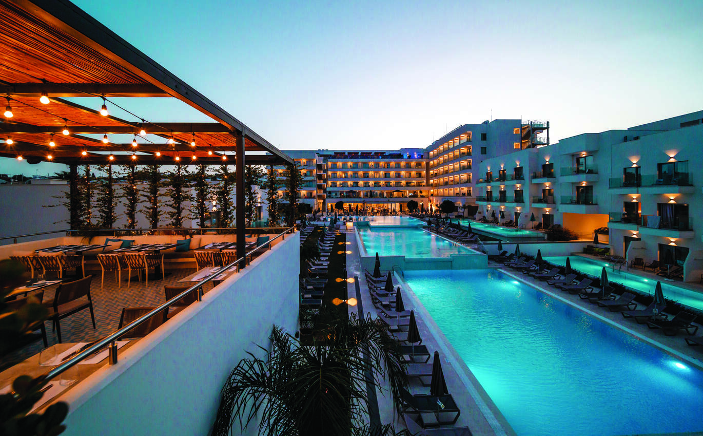 View of the pool area in the dusk with many sun loungers around and the hotel buildings all lit up surrounding. With the undercover restaurant area to the left on a higher level.