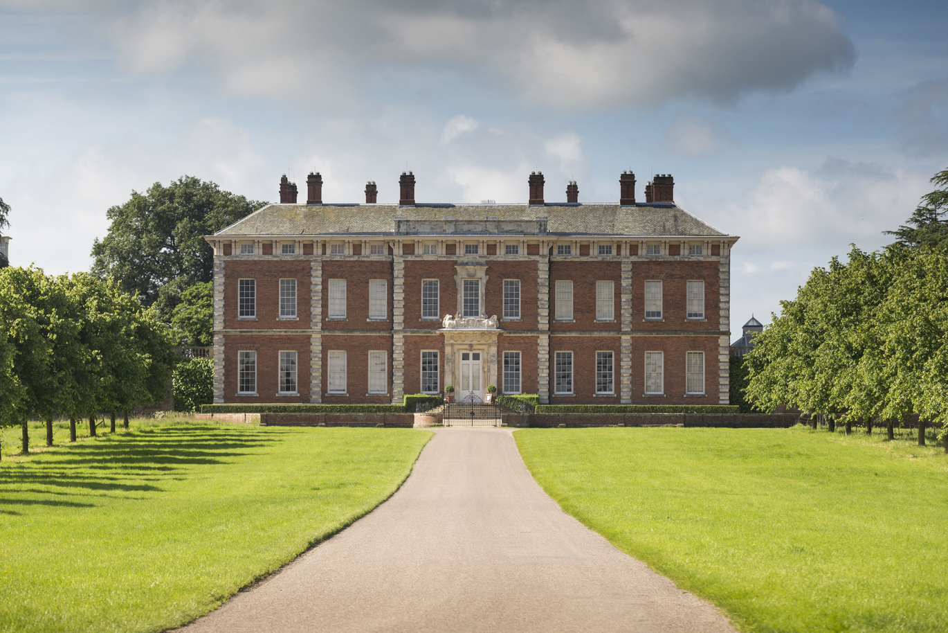 A large red brick building with a straight stone drive leading up to black entrance gates with lush green lawn either side of the path. There are rows of trees set back on the lawn running adjacent to the path. 