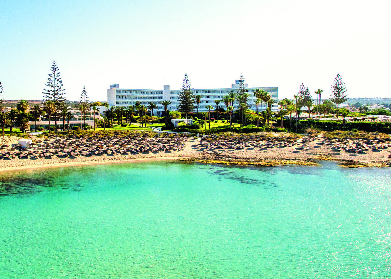 View from above the sea of the resort beach filled with sun loungers and umbrellas with the hotel complex behind, with lush green lawns with palm trees dotted around and the large white hotel building at the back.