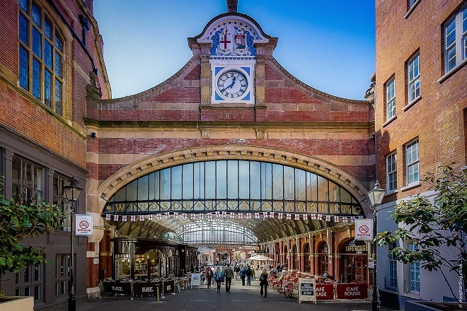 "Photo of entrance to Windsor Royal Station, there are cafes inside either side of the walkway. There is a clock at the top of the entrance with crests above."