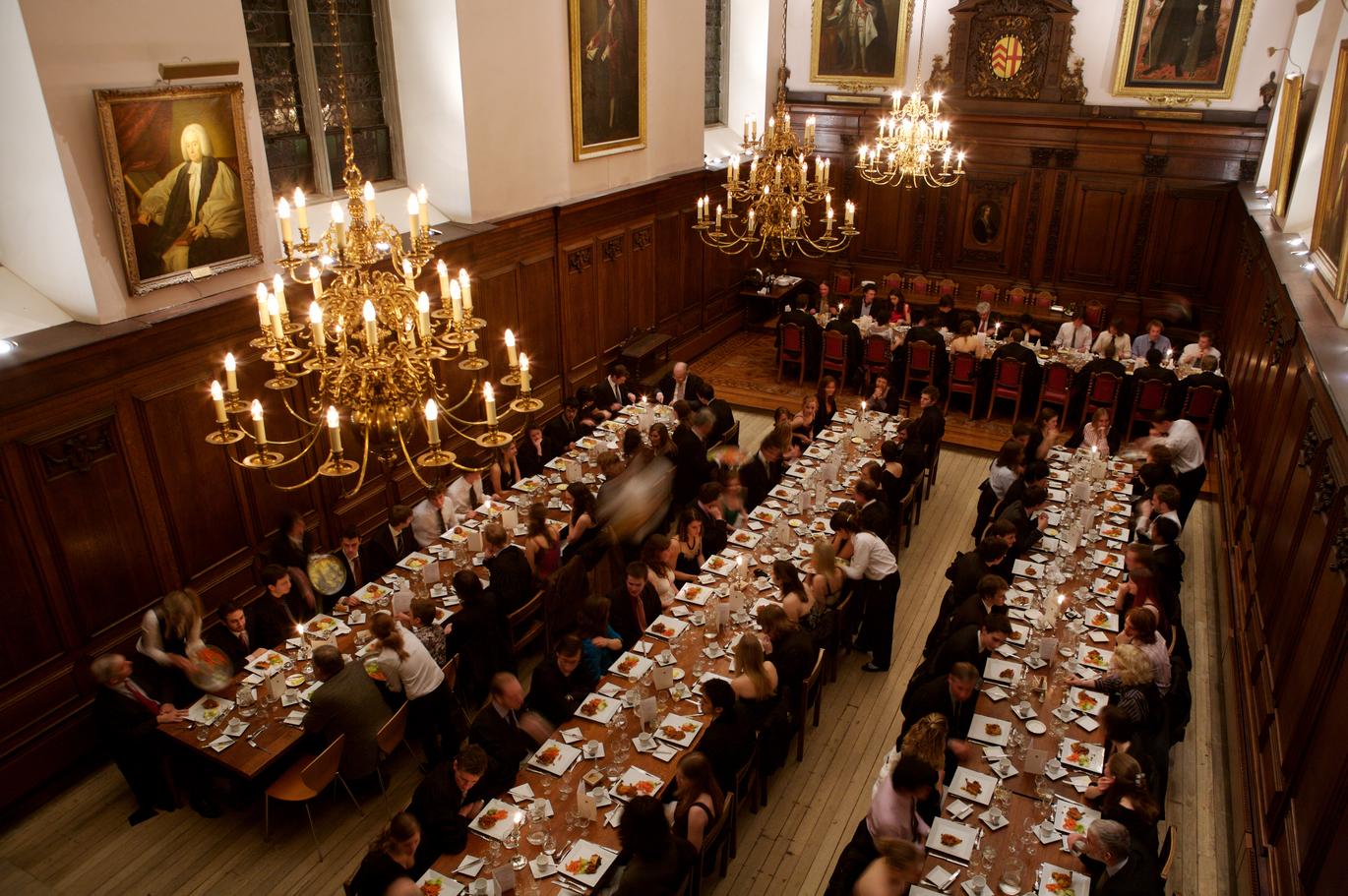 "Birds eye view of formall hall with people seated at long bench tables"