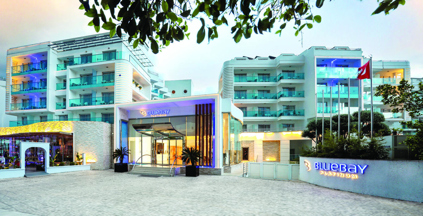 View of the hotel entrance with clusters of hotel rooms and balconies visible behind and a bar/restaurant area to the left of the entrance. The hotel name is lit up on a wall to the right of the entrance 