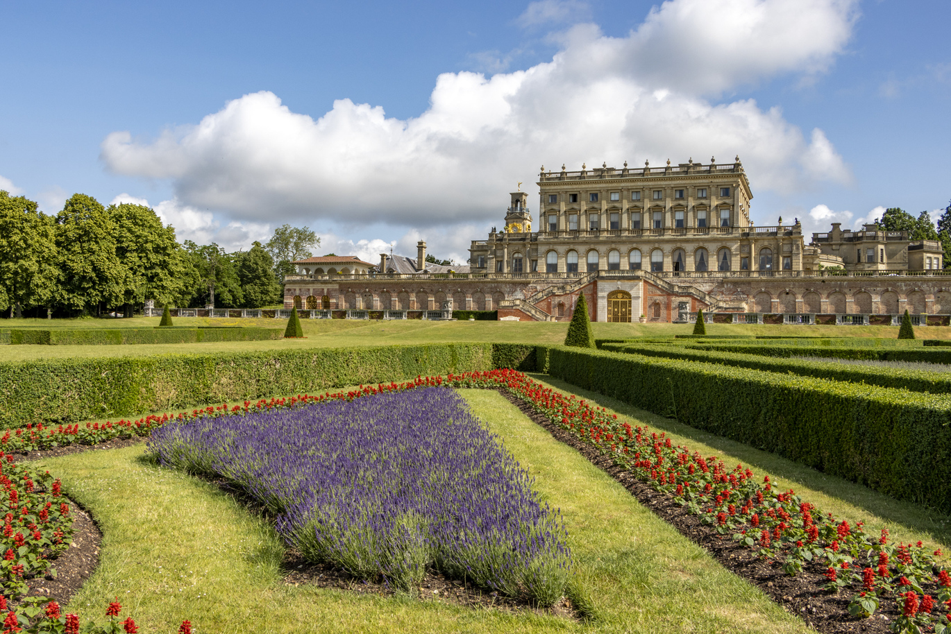 A very, large and magnificent building with a beautiful lush green lawn out the front filled with neatly trimmed hedges in different sizes and shapes with colourful flower beds within the hedges.