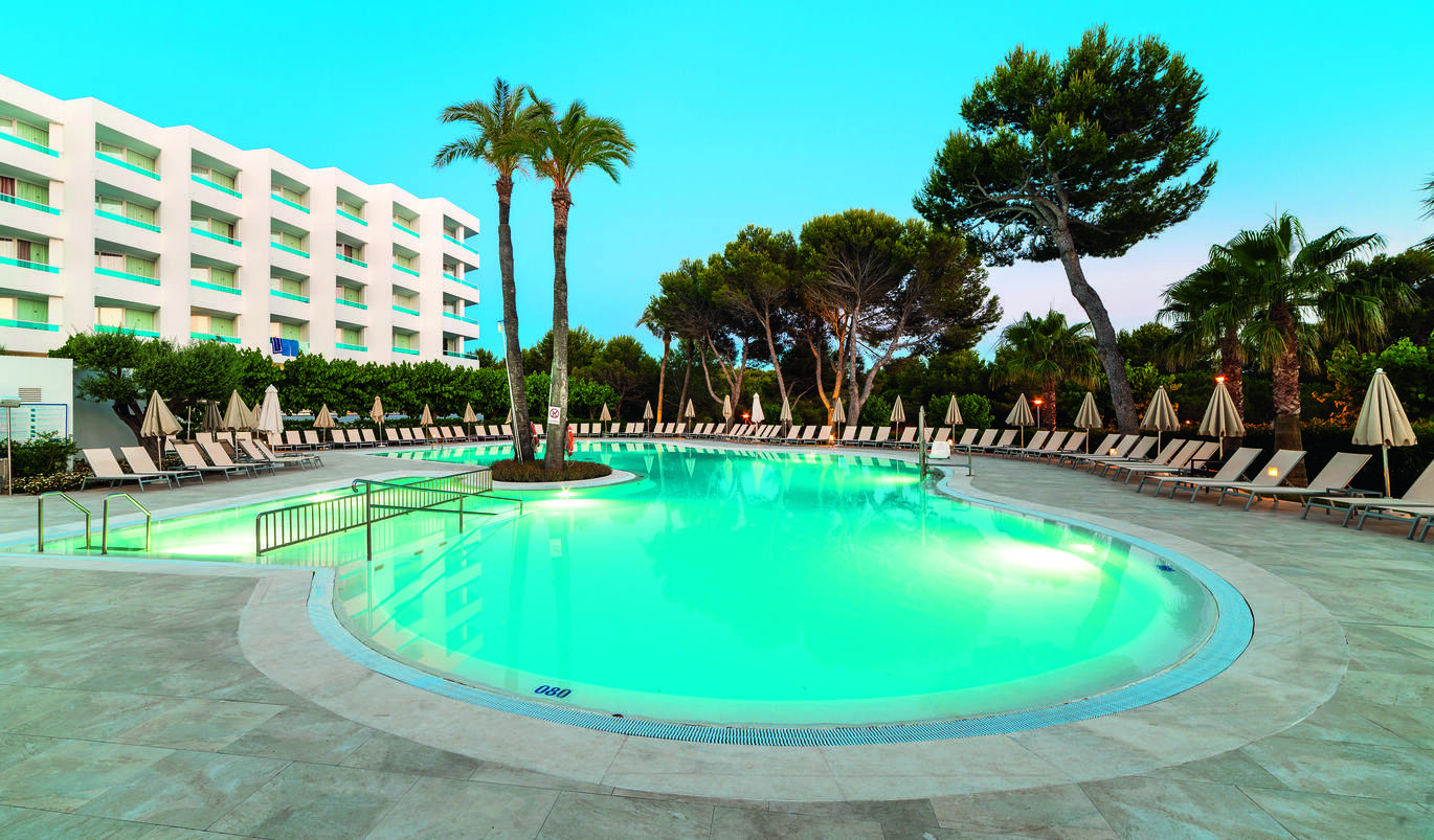 A view in the evening of the small, blue pool with 2 palm trees right at one edge. The pool is surrounded by sandy coloured sun lounders and collapsed umbrellas. Behind this is a large cluster of palm trees and bushes with a view of the hotel building to the left. The sky above is clear and blue.